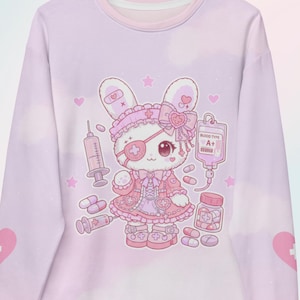 May include: A light purple long-sleeve sweatshirt featuring a central graphic of a white cartoon rabbit wearing a pink nurse-style outfit, an eye patch, and a bow. The design includes medical elements like a syringe, IV bag labeled A+, and pills.