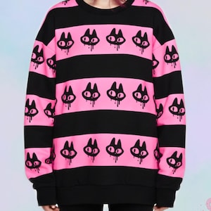 Könnte beinhalten: Schwarz-rosa gestreiftes Sweatshirt mit einem sich wiederholenden Katzenmuster. Das Sweatshirt mit Rundhalsausschnitt hat lange Ärmel. Die Katzenmotive sind schwarz mit tropfenden Details, auf rosa Streifen.