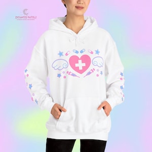 Pode incluir: Moletom branco com um cora&ccedil;&atilde;o rosa, cruz, asas e gr&aacute;ficos com tema m&eacute;dico. O design inclui estrelas, p&iacute;lulas e seringas. As mangas apresentam padr&otilde;es de estrelas em azul e rosa. O texto "ENCHANTED PASTELS" &eacute; vis&iacute;vel no canto superior esquerdo.