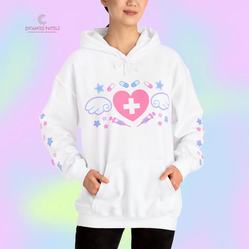 Pode incluir: Moletom branco com um cora&ccedil;&atilde;o rosa, cruz, asas e gr&aacute;ficos com tema m&eacute;dico. O design inclui estrelas, p&iacute;lulas e seringas. As mangas apresentam padr&otilde;es de estrelas em azul e rosa. O texto "ENCHANTED PASTELS" &eacute; vis&iacute;vel no canto superior esquerdo.