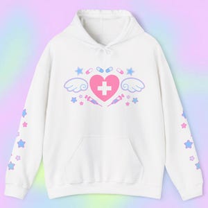 Pode incluir: Moletom branco com bolso frontal e capuz com cord&atilde;o. A frente apresenta um cora&ccedil;&atilde;o rosa com uma cruz branca, asas, estrelas, p&iacute;lulas e seringas. As mangas t&ecirc;m estrelas azuis e rosas.