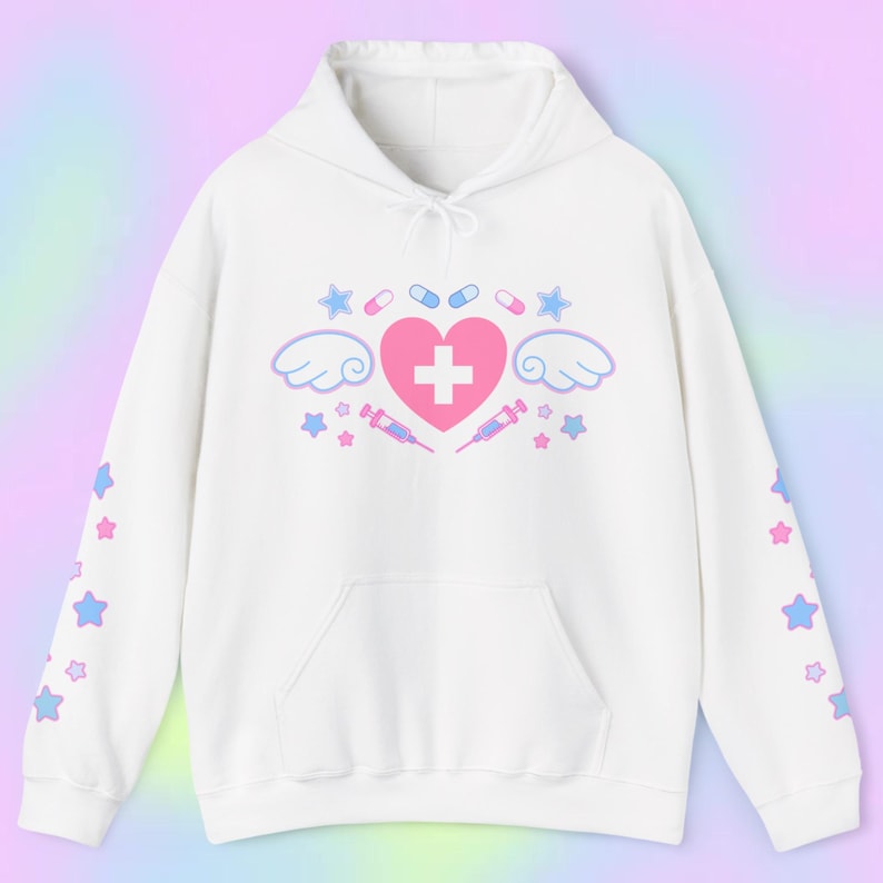 Pode incluir: Moletom branco com bolso frontal e capuz com cord&atilde;o. A frente apresenta um cora&ccedil;&atilde;o rosa com uma cruz branca, asas, estrelas, p&iacute;lulas e seringas. As mangas t&ecirc;m estrelas azuis e rosas.