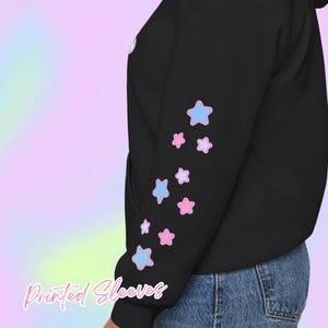 Pode incluir: Moletom preto com estampa de estrelas azul claro e rosa na manga. O texto "Printed Sleeves" est&aacute; escrito em it&aacute;lico na parte inferior da imagem. O fundo &eacute; um gradiente pastel.