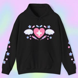 Pode incluir: Moletom preto com um cora&ccedil;&atilde;o rosa, uma cruz branca, asas, estrelas, p&iacute;lulas e seringas. As mangas t&ecirc;m estrelas azuis e rosas. O moletom tem capuz e um bolso frontal.