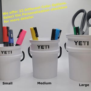 May include: Three white Yeti brand containers with black lettering, labeled "Small", "Medium", and "Large". Each container holds various writing utensils, including markers and pens.