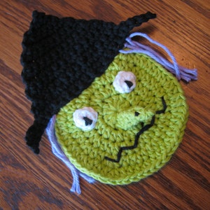 May include: A green crocheted witch face with a black crocheted hat and purple yarn hair. The witch has a crooked nose and two white eyes.