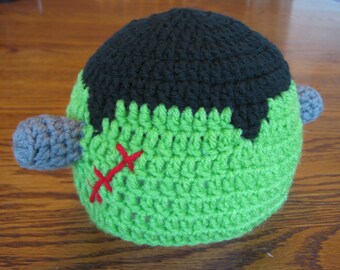 May include: Green and black crocheted hat with grey yarn accents. The hat has a stitched red line design on the front.