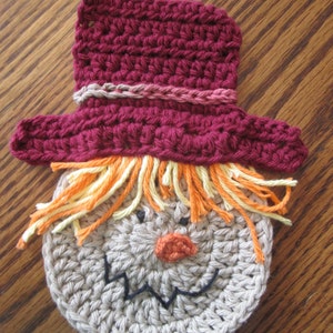 Puede incluir: Una cara de espantapájaros de ganchillo con un sombrero burdeos, pelo naranja y amarillo y una sonrisa cosida negra.