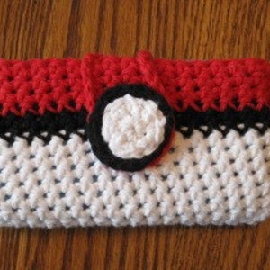 Op de afbeelding: Een gehaakte telefoonhoes in Pokeball-thema met een rode, zwarte en witte kleurstelling. De hoes heeft een kleine zwarte knop in het midden.