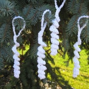 Puede incluir: Adornos de hielo de crochet blanco colgados de una rama de pino verde. Los carámbanos están hechos con un punto retorcido y tienen un aspecto texturizado.