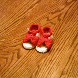 K&ouml;nnte beinhalten: Ein Paar geh&auml;kelte Babyschuhe in Wei&szlig; mit roten Schleifen. Die Schuhe liegen auf einer Holzoberfl&auml;che.