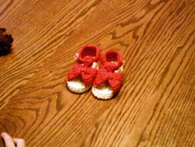 K&ouml;nnte beinhalten: Ein Paar geh&auml;kelte Babyschuhe in Wei&szlig; mit roten Schleifen. Die Schuhe liegen auf einer Holzoberfl&auml;che.