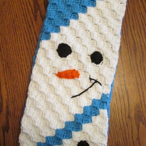 May include: A white crocheted scarf with a blue border and a snowman face. The snowman has black eyes, an orange nose, and a black smile.