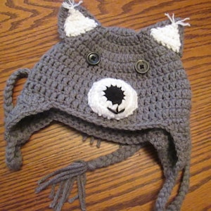 Puede incluir: Sombrero de crochet gris con orejas blancas y una cara blanca con ojos y nariz negros. El sombrero tiene una larga borla gris.