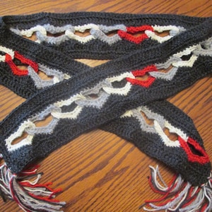 May include: A black crocheted scarf with white, gray, and red accents. The scarf has a fringe on both ends and features a repeating heart pattern.