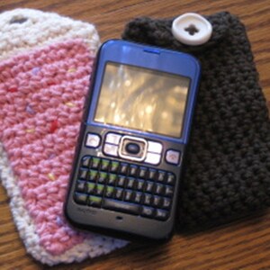 May include: A blue cell phone with a black keyboard and a white button is in a pink and white crocheted phone case. The phone case is next to a brown crocheted phone case.