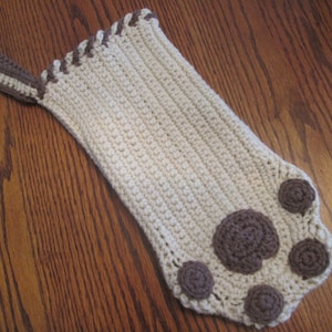 May include: A crocheted stocking with a brown paw print design. The stocking is cream colored with brown trim.