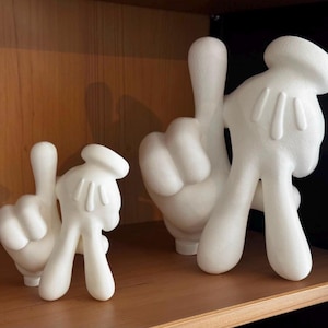 Op de afbeelding: Twee witte, cartoonachtige handsculpturen in verschillende formaten, elk met vier vingers, een duim en drie duidelijke lijnen op de rug van de hand. De handen zijn gepositioneerd met de wijsvinger omhoog en de andere vingers naar binnen gekruld.
