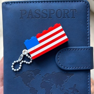 May include: A navy blue passport holder with the word "PASSPORT" embossed on the front. A small charm of the American flag made of toy blocks is attached. The holder features a world map design and a snap closure.
