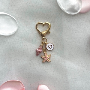 May include: A gold-toned heart-shaped lobster clasp keychain featuring three dangling charms: a small pink seashell, a pink starfish, and a circular white charm with the letter D printed in black, all connected by small gold jump rings.