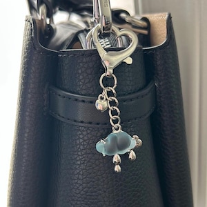 May include: A black leather handbag with a silver-tone metal clasp and a cloud charm. The charm features a light blue cloud with silver raindrops and a small heart. The handbag has a top handle and a shoulder strap.