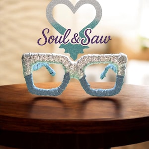 Puede incluir: Gafas decorativas con un diseño plateado y azul. Las monturas están adornadas con purpurina y tela, y las lentes están abiertas. La imagen también presenta una decoración en forma de corazón y el texto "Soul & Saw".