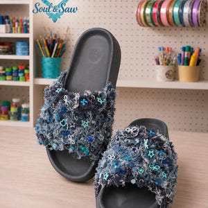 May include: A pair of dark gray slide sandals featuring wide straps covered in frayed blue denim fabric. The denim is embellished with various small blue and turquoise charms, including stars, flowers, bows, and beads, creating a textured look.