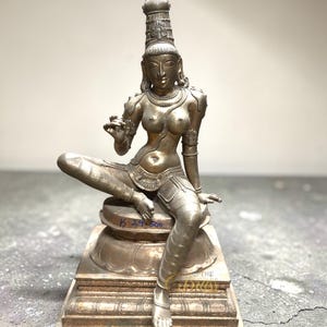 May include: A detailed brass sculpture of a seated female figure, adorned with intricate jewelry and a tall headdress. The figure is posed on a decorative base, showcasing fine craftsmanship. The sculpture has a metallic finish and is likely a religious or cultural artifact.