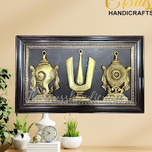May include: A framed wall decor with three gold-colored metal religious symbols. The symbols are a conch shell, a trident, and a wheel. The frame is black with a gold trim.