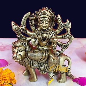 Può includere: Una statua in ottone dorato di una divinità indù, Durga, che cavalca un leone. La statua è dettagliata con intricate sculture e ha più braccia. La statua è seduta su una superficie bianca con petali di fiori gialli e rosa.