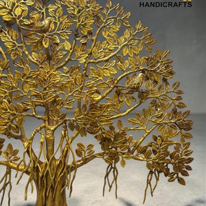 May include: A gold-coloured metal tree sculpture with intricate leaf and branch detailing, with two small birds nestled in the leaves. The sculpture has a textured finish and the text "THE Brass HANDICRAFTS" in gold.