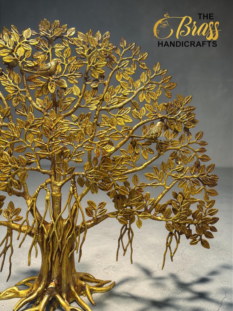 May include: A gold-coloured metal tree sculpture with intricate leaf and branch detailing, with two small birds nestled in the leaves. The sculpture has a textured finish and the text "THE Brass HANDICRAFTS" in gold.