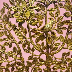 May include: A gold-coloured metal tree sculpture featuring detailed leaves and a perched bird. The branches and leaves are intricately designed, creating a naturalistic aesthetic. The background is a soft pink, enhancing the sculpture's warm tones.