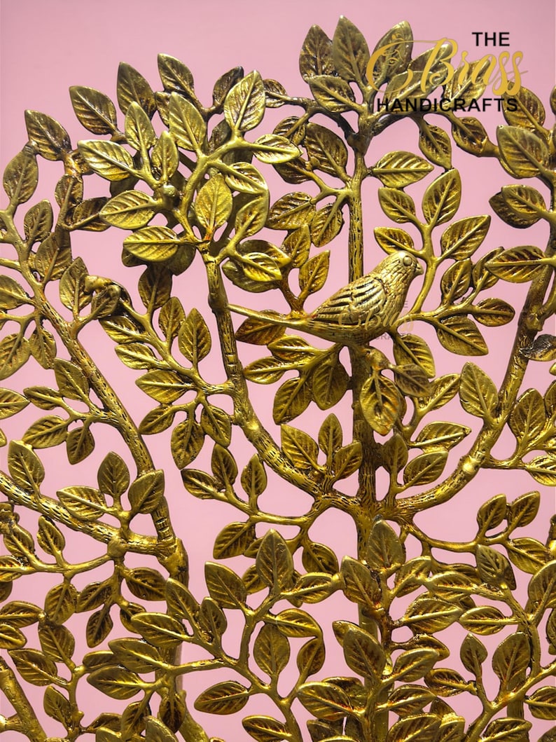 May include: A gold-coloured metal tree sculpture featuring detailed leaves and a perched bird. The branches and leaves are intricately designed, creating a naturalistic aesthetic. The background is a soft pink, enhancing the sculpture's warm tones.