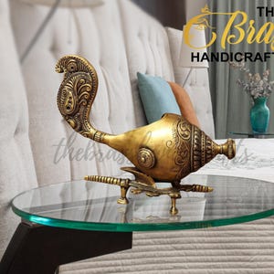 May include: A gold-colored brass peacock-shaped oil lamp with intricate carvings. The lamp is sitting on a glass table top.