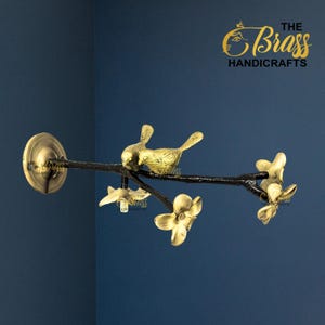 Op de afbeelding: Een messing wanddecoratie met een tak met twee vogels en bloemdetails. De tak is zwart en de vogels en bloemen zijn goudkleurig. Het logo The Brass Handicrafts staat rechtsboven.