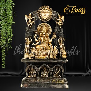 Pode incluir: Uma estátua dourada de uma deusa hindu sentada em um trono de lótus. A deusa tem quatro braços e está adornada com joias. A estátua é cercada por figuras menores e é colocada sobre um fundo preto. O texto "The Brass Handicrafts" é visível no canto superior direito.