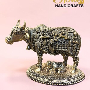 Może przedstawiać: Szczegółowa rzeźba z mosiądzu przedstawiająca krowę z misternymi rzeźbami, stojącą na ozdobnej podstawie. Rzeźba zawiera mniejsze cielę i napis "THE Brass HANDICRAFTS". Krowa jest w kolorze złotym.
