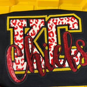 May include: A black t-shirt featuring a large applique design with the letters KC in a white and red leopard print pattern outlined in yellow. Overlaid is the word Chiefs in red cursive script, all embellished with shimmering sequins.
