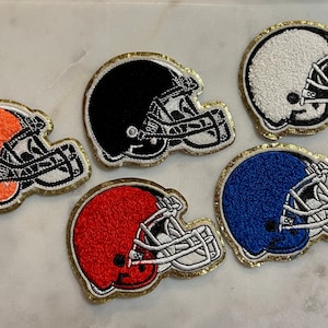 May include: Five embroidered football helmet patches in orange, black, white, red, and blue. Each helmet has a gold glitter border and a black facemask. These patches are suitable for sports-themed crafts.