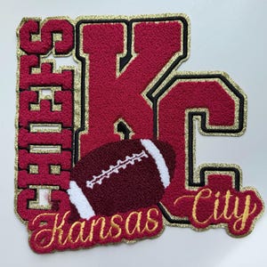 May include: A decorative patch featuring the text Chiefs and Kansas City in red chenille with gold glitter borders. Large red chenille letters KC are centered behind a dark red football with white laces. The design has a black outline throughout.
