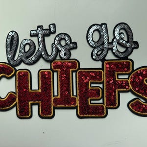 May include: A decorative patch featuring the text "let's go" in silver sequins above the word "CHIEFS" in deep red sequins with gold trim. The letters are outlined in black, creating a bold, layered appearance for this embroidered applique.