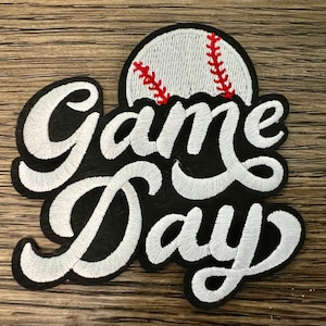 May include: White embroidered patch with the words "Game Day" in a cursive font. A white baseball with red stitching is above the words.