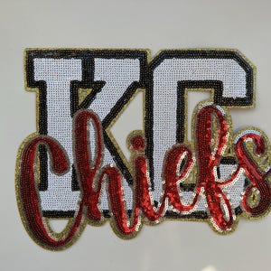 May include: A decorative patch featuring the letters KC in white sequins with black and gold borders, overlaid with the word Chiefs in red cursive script sequins. The entire design is outlined in gold sequins against a plain white background.