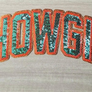May include: A sequined patch in the shape of the word "SHOWGIRL". The letters are filled with teal sequins and outlined in orange and black sequins. The patch is arched and sits on a light wood grain surface.