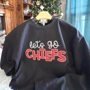 May include: Black crewneck sweatshirt with the text "let's go CHIEFS" in white and red glitter lettering. The sweatshirt is displayed on a wooden hanger. The background includes a Christmas tree and a wooden cabinet.