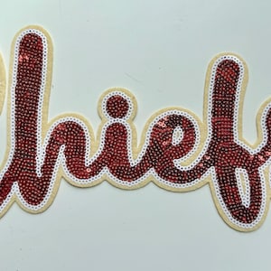 May include: A decorative patch featuring the word Chiefs in cursive script. The lettering is filled with dark red sequins and outlined with a border of white sequins, all set against a light beige felt backing material.