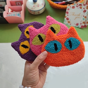 May include: Three colourful felt cat head ornaments with embroidered eyes. The cat heads are purple, pink, and orange. The eyes are yellow, green, and blue.