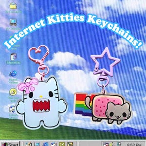 May include: Two acrylic keychains with the text "Internet Kitties Keychains!". One keychain is a white cat with a pink bow and heart-shaped clasp. The other is a pink cat with a rainbow tail and a star-shaped clasp.