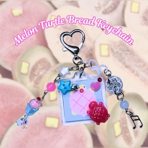 May include: A pink and blue turtle bread keychain with a heart-shaped silver clasp. The keychain has a variety of charms, including a star, a button, a pink heart, a silver clover, and a blue music note.